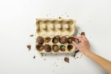 Kadın eli, çikolata yumurtalarını ve çok renkli şeker süslemelerini açık arkaplandaki tepsiden alır ya da tutar. Paskalya konsepti, paskalya hediyeleri, tavşancık. Pankart. Düz yatış, üst görünüm.