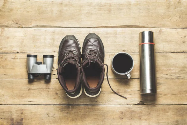 Boots for trail, binoculars, thermos, cup with tea on a wooden background. The concept of hiking, tourism, camp, mountains, forest. Banner. Flat lay, top view.