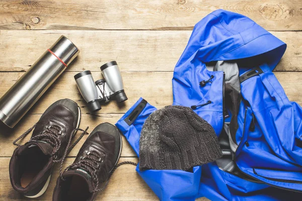 Hiking boots, jacket, binoculars, shirt, hat, backpack on a wooden background. The concept of hiking, tourism, camp, mountains, forest. Banner. Flat lay, top view.