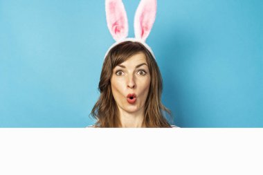 Tavşan kulaklı ve samimi duyguları olan neşeli, genç esmer kadın mavi arka plandaki kartondan dışarı bakıyor. Paskalya konsepti, sürpriz, saklambaç, duygusal. Yüz ifadesi. Pankart.