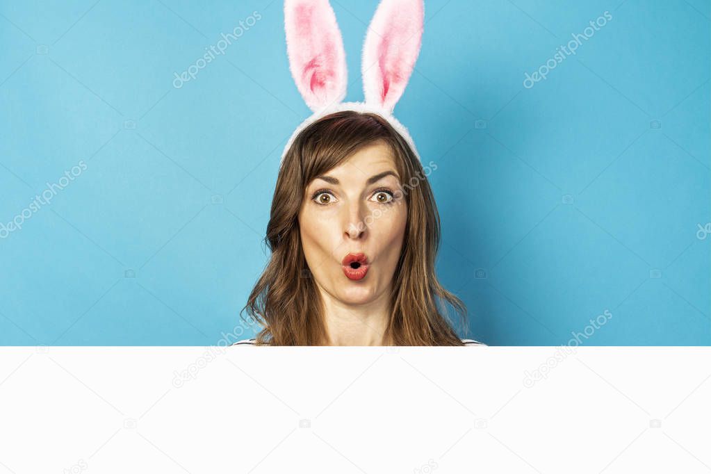 Joyful Young brunette woman with bunny ears and sincere feelings peeks out from behind cardboard on a blue background. Easter concept, surprise, hide and seek, emotional. Face expression. Banner.