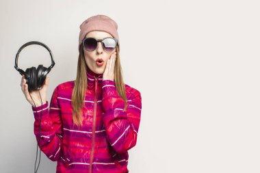 Arkadaş canlısı duygusal. Gözlüklü, şapkalı ve pembe spor ceketli genç bir kadın, fotokopi alanı olan beyaz bir arka planda kulaklık tutuyor. Modern moda müzik konsepti. Yüz ifadesi. Pankart.