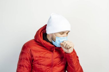 Yüzünde tıbbi maske olan genç bir adam hafif bir arka planda öksürüyor. Soğuk algınlığı, virüs, bulaşıcı hastalıklar.