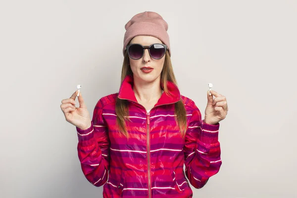 Friendly Young woman in glasses, hat and pink sports jacket with a smiley face holds wireless headphones on a white background. Concept modern style, cool music. Face expression. Banner.