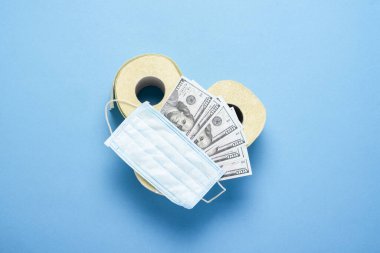 Tuvalet kağıdı ruloları, tıbbi maske ve mavi arka planda dolar banknotları. Düz yerde, üst tarafta. Konsept hijyen ürünü, virüs, eksiklik, karantina.