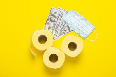 Tuvalet kağıdı ruloları, tıbbi maske ve sarı arka planda dolar banknotları. Düz yerde, üst tarafta. Konsept hijyen ürünü, virüs, eksiklik, karantina.