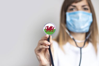 Tıbbi maskeli kadın doktor arka planda steteskop tutuyor. Galler bayrağı eklendi. Tıp, ilaç seviyesi, virüs, salgın hastalık.