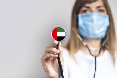 Tıbbi maskeli kadın doktor arka planda steteskop tutuyor. Birleşik Arap Emirliği bayrağı eklendi. Tıp, ilaç seviyesi, virüs, salgın hastalık.
