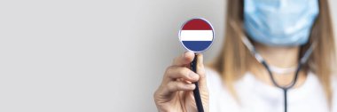 Tıbbi maskeli kadın doktor arka planda steteskop tutuyor. Hollanda bayrağı eklendi. Tıp, ilaç seviyesi, virüs, salgın hastalık. Uyarı