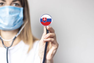 Tıbbi maskeli kadın doktor arka planda steteskop tutuyor. Slovakya bayrağı eklendi. Tıp, ilaç seviyesi, virüs, salgın hastalık.