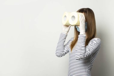 Tıbbi maske takan ve eldiven takan genç bir kız tuvalet kağıdı rulolarını dürbün şeklinde tutuyor. Işık arkaplanındaki dürbünle bakıyor. Karantina, kıtlık ve panik.
