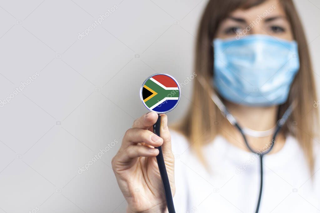 female doctor in a medical mask holds a stethoscope on a light background. Added flag of South Africa. Concept medicine, level of medicine, virus, epidemic.