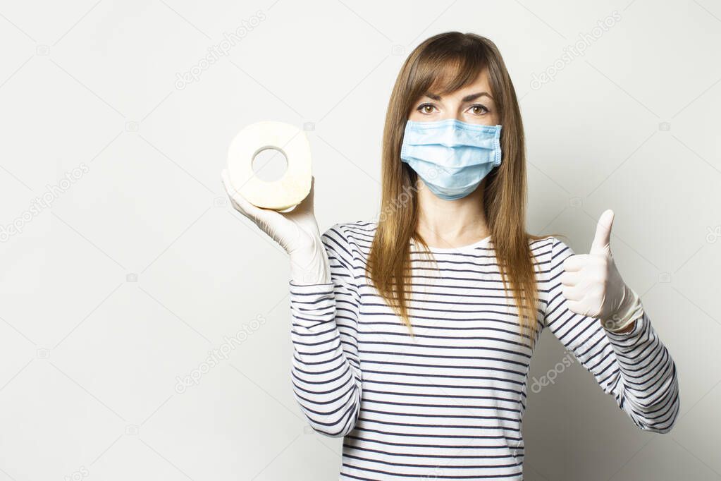young girl in a medical mask and medical gloves holding a roll of toilet paper and shows the gesture class on a light background. Concept quarantine, shortage, panic.