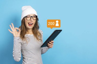 Şapkalı, gözlüklü ve tişörtlü neşeli yüzlü genç bir kadın elinde bir tablet tutuyor ve izole edilmiş mavi arka planda 