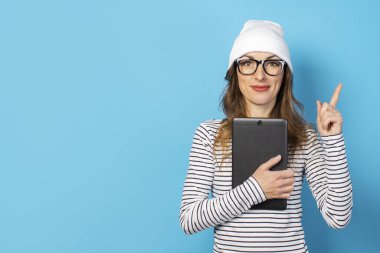 Şapkalı, gözlüklü ve tişörtlü mutlu yüzlü genç bir kadının portresi elinde bir tablet tutuyor ve parmağını izole edilmiş mavi bir arkaplanı işaret ediyor. Duygusal yüz..