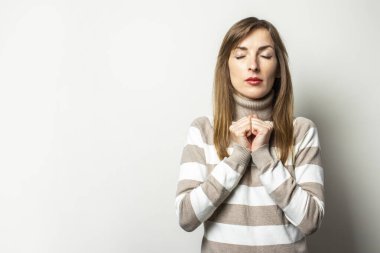 Çizgili süveter giymiş genç bir kadının portresi ellerini göğsüne koydu ve izole bir arka planda gözlerini kapattı. Duygusal bir yüz. Bir şeyin jest beklentisi.
