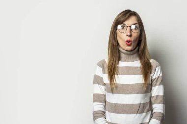Çizgili süveter ve gözlüklü, izole ışık arka planında şaşırmış bir yüze sahip genç bir kadının portresi. Duygusal bir yüz. Şaşırtıcı bir jest..