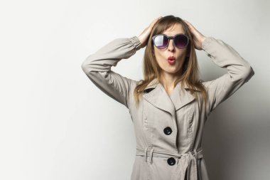 Klasik ceketli ve güneş gözlüklü genç bir kadın aydınlık arka planda şaşırmış bir yüzle ellerini tutuyor. Duygusal bir yüz. Klasik tarz, Fransız kadın, sürpriz, şok.