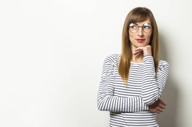 Uzun kollu, gözlüklü, uzun kollu genç bir kadının portresi yan bakıyor ve elini, izole ışık arka planında çenesinin altından tutuyor. Duygusal bir yüz. Düşünmek için jest, hayal kurmak.