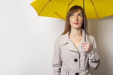Mutlu, neşeli genç bir kadın sarı bir şemsiye tutuyor ve gri bir arka planda ona bakıyor. Kavramsal jestler, yağmur, kötü hava.