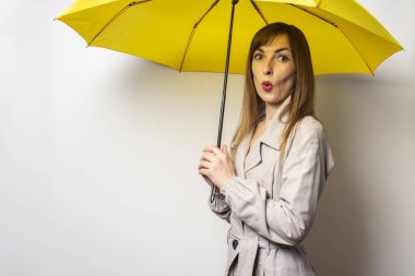Klasik pelerinli genç bir kadın portresi sarı bir şemsiyenin altında şaşırmış bir yüzle izole bir ışık arka planında. Duygusal bir yüz. Kötü hava, yağmur, hava tahmini.