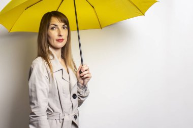 Klasik ceketli, sarı şemsiyeli genç ve dost canlısı bir kadının portresi izole bir ışık arka planına bakar. Duygusal bir yüz. Kötü hava, yağmur, hava tahmini.