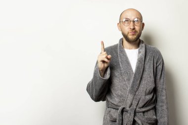 Sabahlık ve gözlüklü sakallı genç kel bir adamın portresi yukarı bakar ve izole bir ışık arka planını işaret eder. Duygusal yüz..