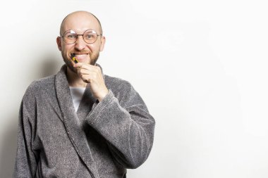 Bornozlu ve gözlüklü sakallı genç bir adamın portresi izole bir arka planda dişlerini fırçalıyor. Oral bakım konsepti, kişisel hijyen.