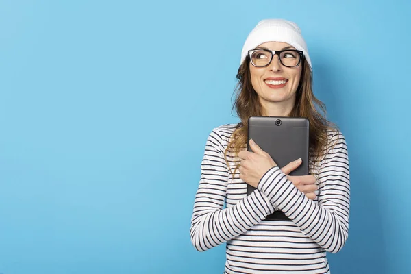 Portrait of a young friendly woman in a hat, glasses and a T-shirt holding a tablet in her hands and looking aside on an isolated blue background. Emotional face. Social network concept, blogger.