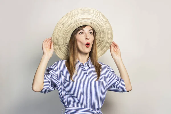 Portrait of a young friendly woman with a surprised face in a dress and a wide-brimmed hat on an isolated light background. Emotional face. Gesture of surprise, joy.