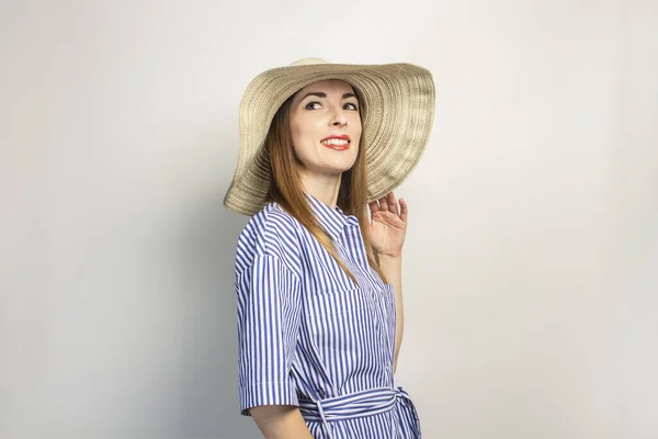 Portrait of a young friendly woman with a smile in a dress and a wide-brimmed hat on an isolated light background. The hat covers part of the face. Emotional face. A gesture of joy. Banner.