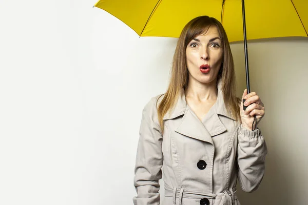 Portrait Young friendly woman in a classic cloak with a surprised face under a yellow umbrella on an isolated light background. Emotional face. Bad weather, rain, weather forecast.