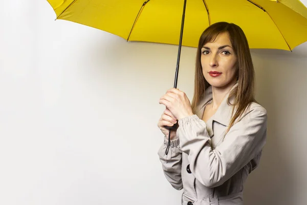 Portrait of a young friendly woman in a classic jacket under a yellow umbrella on an isolated light background. Emotional face. Bad weather, rain, weather forecast.