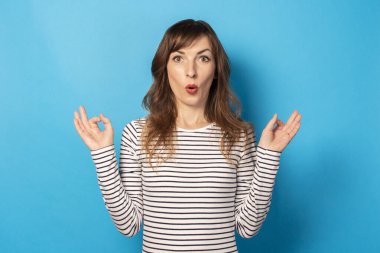 Sıradan bir tişörtle şaşırmış genç bir kadının portresi izole edilmiş mavi bir arka planda zen hareketi yapıyor. Duygusal bir yüz. Rahatlama, meditasyon, stres kavramı.