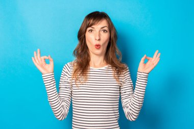 Sıradan bir tişörtle şaşırmış genç bir kadının portresi izole edilmiş mavi bir arka planda zen hareketi yapıyor. Duygusal bir yüz. Rahatlama, meditasyon, stres kavramı.