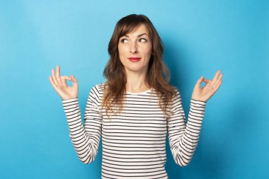 Sıradan bir tişörtle gülümseyen genç bir kadının portresi zen jesti yapıyor ve izole edilmiş mavi arka planda kıça bakıyor. Duygusal bir yüz. Rahatlama, meditasyon, stres kavramı.