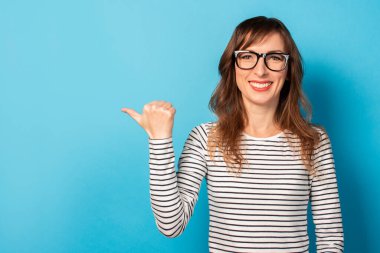 Sıradan bir tişört ve gözlük takmış gülümseyen genç bir kadının portresi izole bir arka planda bir parmağı işaret ediyor. Duygusal bir yüz. Şuna bak, dikkat et..