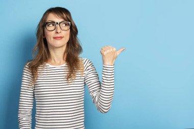 Gözlüklü ve çizgili süveter giyen sevimli genç bir kadın mavi arka planda başparmağını yana gösteriyor. Duygusal bir yüz. Şaşırdın mı, dikkatini ver..
