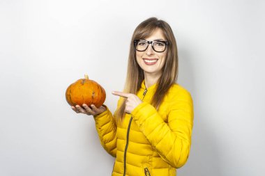 Sarı ceketli genç kız bir balkabağı tutuyor ve arka planda onu işaret ediyor. Cadılar Bayramı konsepti, sonbahar, kutlama.