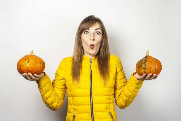 shocked young girl in a yellow jacket holds two pumpkins on a light background. Halloween concept, autumn, celebration.