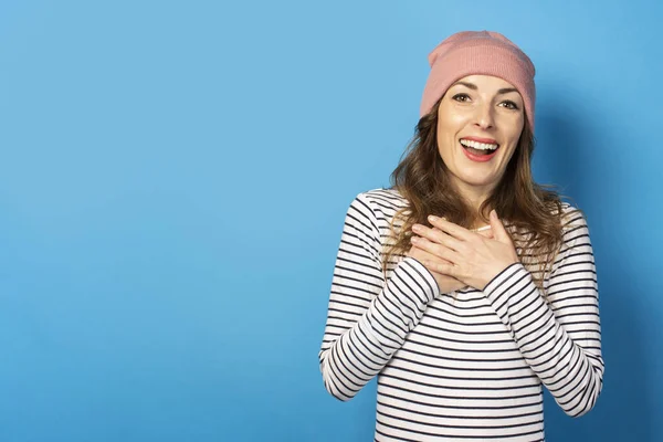 Cute young woman with a surprised face in a hat and striped sweater laid two palms on her chest against a blue background. Emotional face. Gesture of excitement, joy, surprise.