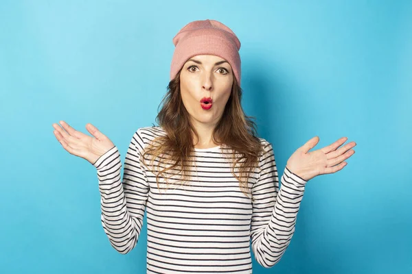 Cute young woman with a surprised face in a hat and striped sweater shrugs her hands on a blue background. Emotional face. A gesture of bewilderment, surprise, disappointment.