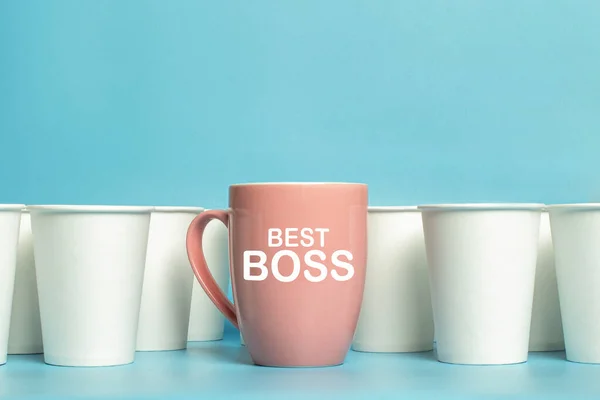 Pink cup surrounded by white paper cups on a blue background. Concept boss, unique, friendly team, feminism. Copy space. Added text Best Boss.