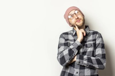 Şapkalı, ekose gömlekli ve gözlüklü genç bir adamın portresi izole bir arka planda görünüyor. Duygusal bir yüz. Rüya jesti planlaması.