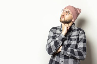 Şapkalı, ekose gömlekli ve gözlüklü genç bir adamın portresi izole bir arka planda görünüyor. Duygusal bir yüz. Rüya jesti planlaması.