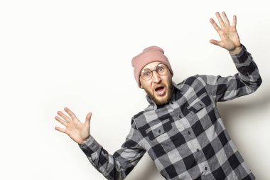 Şapkalı, ekose gömlekli ve gözlüklü genç bir adamın portresi, yalıtılmış ışık arka planında şaşırmış bir yüzle yan tarafından dışarı bakıyor. Duygusal bir yüz. Şaşırtıcı bir jest..