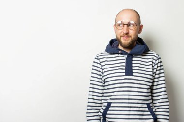 Kapüşonlu gözlüklü sakallı kel bir gencin portresi izole edilmiş bir ışık arka planına bakıyor. Duygusal yüz..