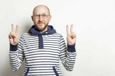 Kapüşonlu ceket gözlüklü kel bir gencin portresi parmaklarıyla jest yapıyor. Tamam, her şey yolunda, selamlar, izole edilmiş ışık arka planında barış. Duygusal yüz..