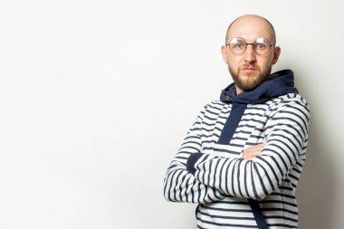 Kel bir genç adamın portresi, sakallı, gözlüklü, kapüşonlu bir süveter giymiş, kollarını göğsüne kavuşturmuş, izole bir arka planda. Duygusal bir yüz. Konsantre hareket..