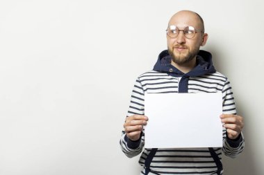Gözlüklü kel bir genç adamın portresi, kapüşonlu bir süveter önünde boş bir kağıt tutuyordu, izole bir arka planda yan tarafa bakıyordu. Boşluğu kopyala.